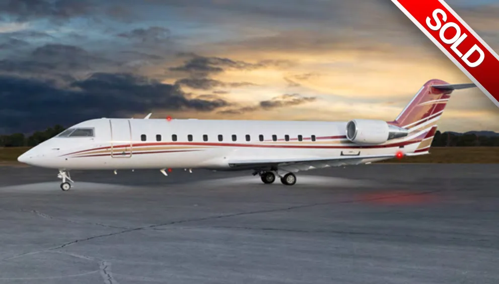 A white business jet with red and gold stripes is parked on a tarmac at sunset.