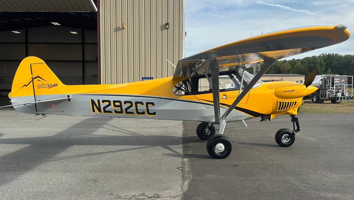 A yellow and gray NX Cub aircraft, N292CC, parked on an asphalt tarmac in front of a hangar.