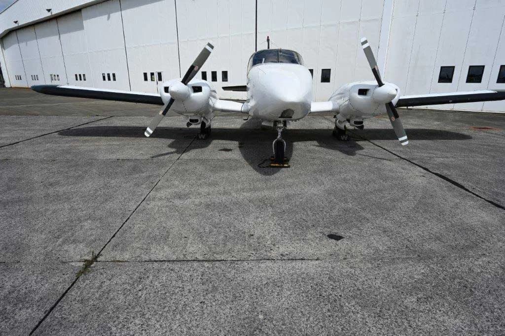 A white twin-engine propeller aircraft with black propellers, viewed from the front, parked on a concrete apron in front of a white hangar.