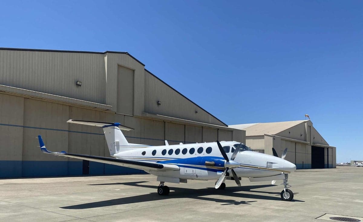 A front-quarter view of the Beechcraft King Air 350 with blue and white livery, parked in front of hangars.