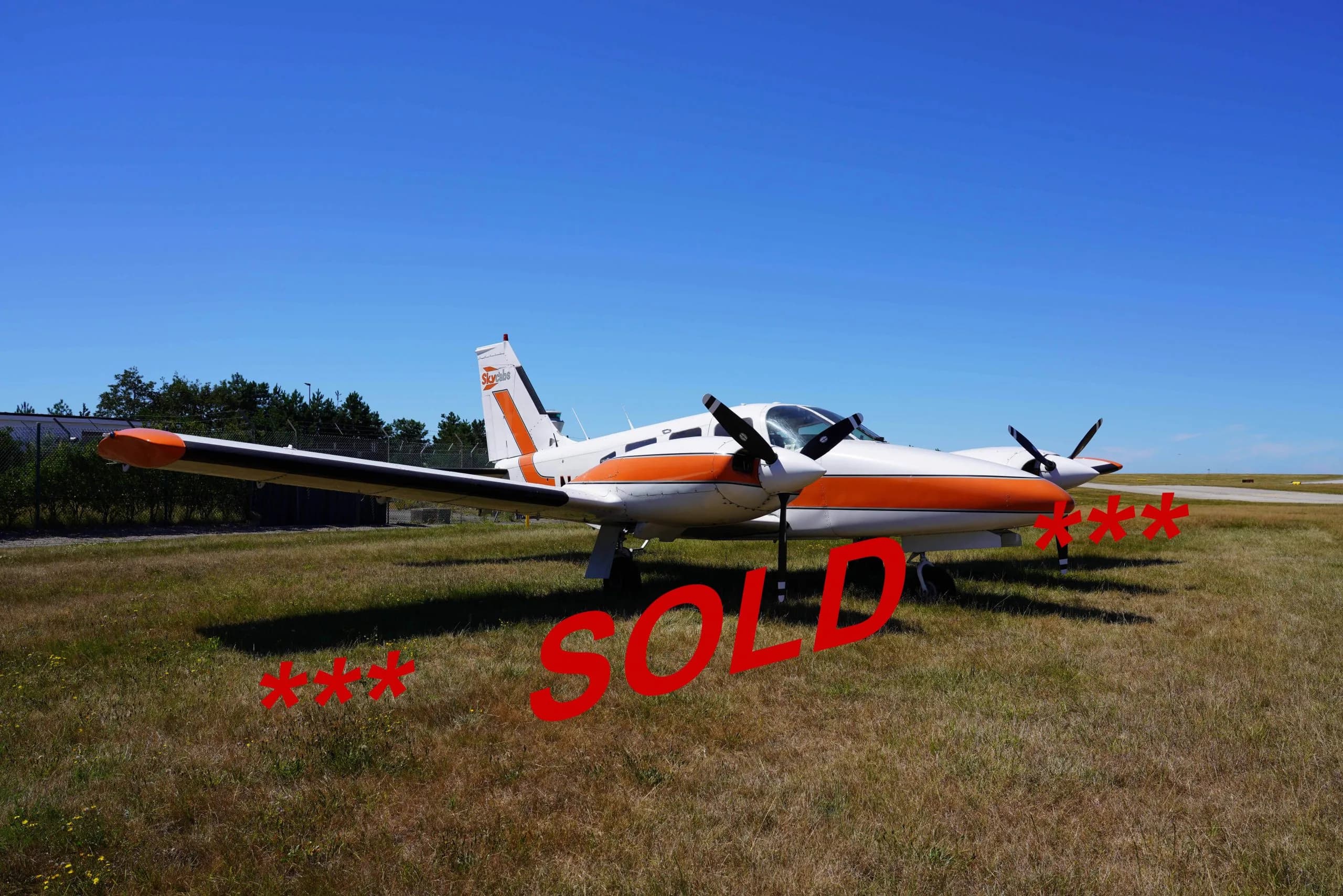 A white twin-engine propeller aircraft with orange stripes and 'Skycabs' logo on the tail, parked on a grassy field under a clear blue sky.