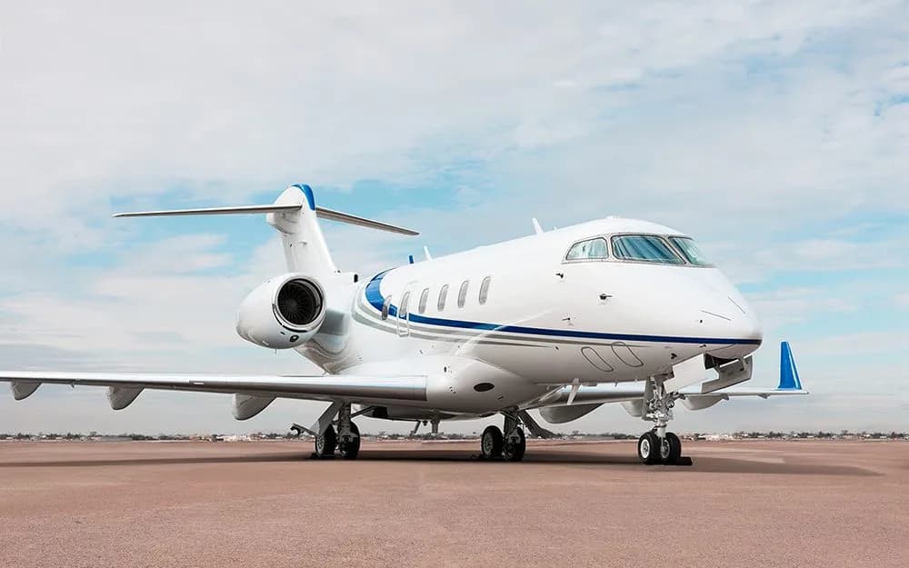 A white private jet with blue accent stripes is parked on an airfield under a partly cloudy sky.