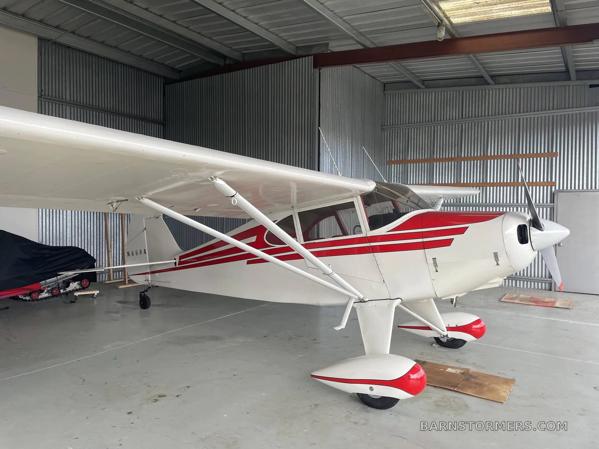 A white and red Piper PA-12 Super Cruiser aircraft is parked inside a hangar.