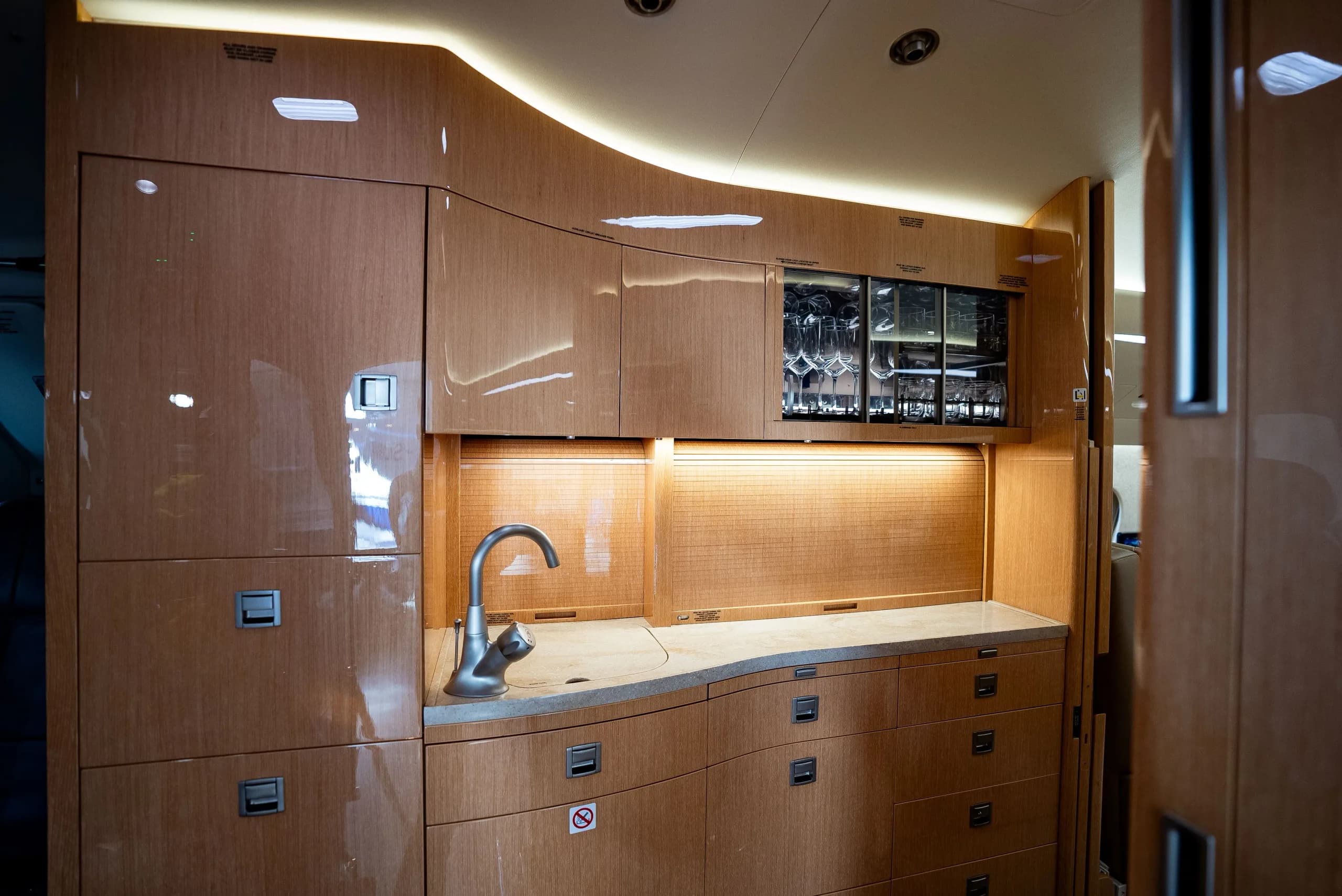 The galley area of a private jet, featuring wooden cabinetry, a sink, and a display cabinet with glassware.