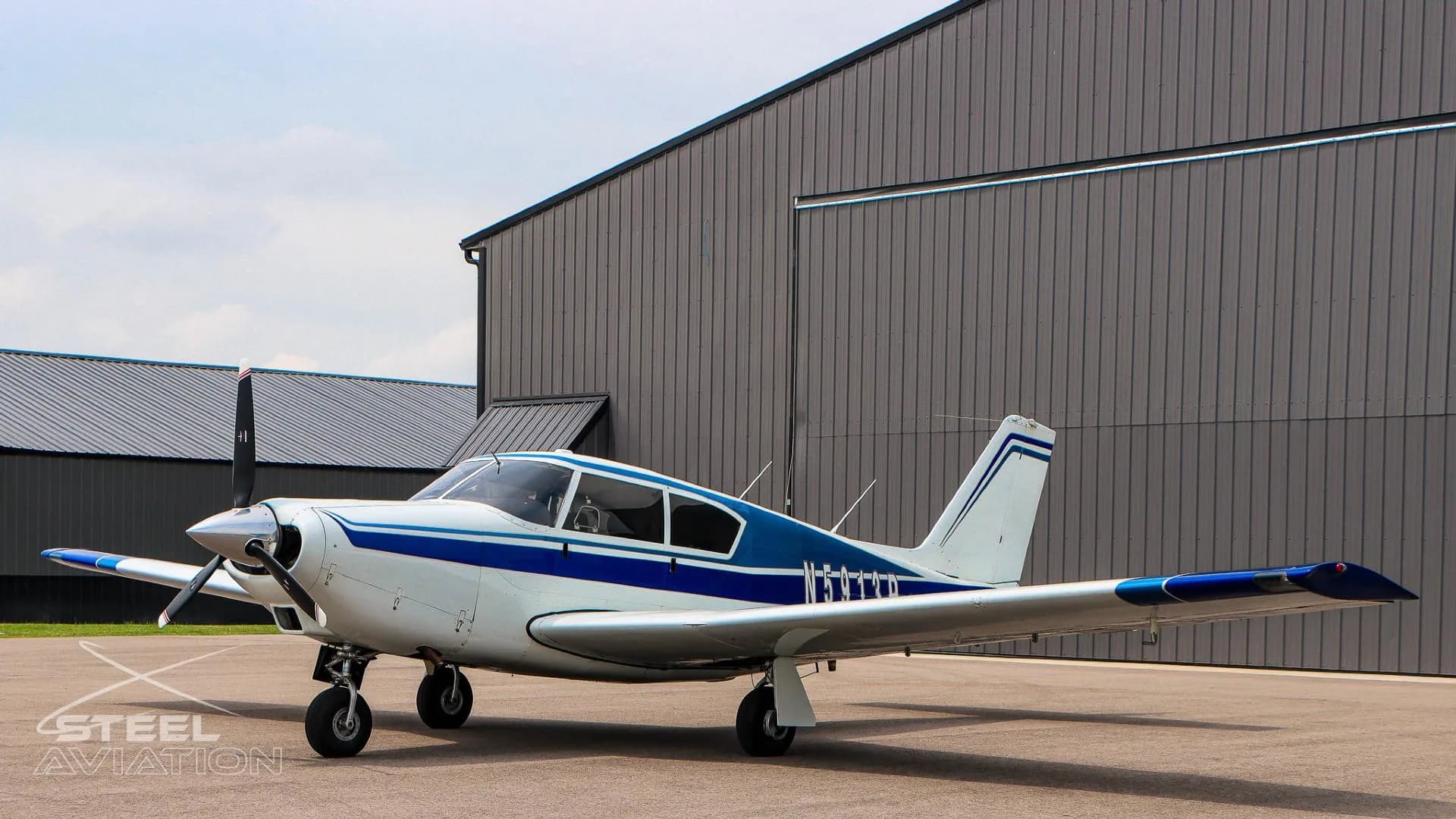 A side view of a white and blue single-engine aircraft parked in front of a dark gray hangar.