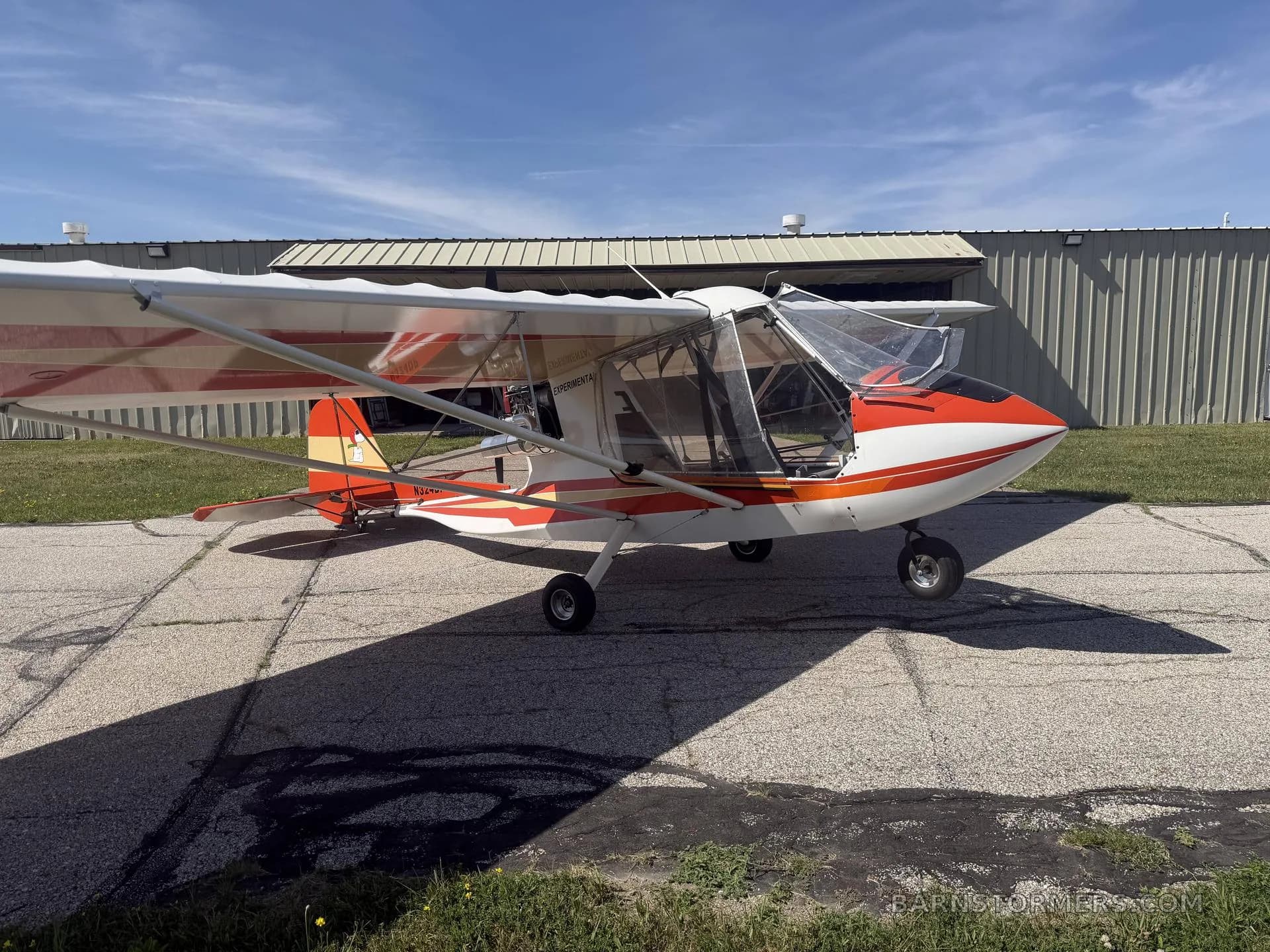 A white and orange Challenger ultralight aircraft parked on an asphalt apron in front of a hangar.