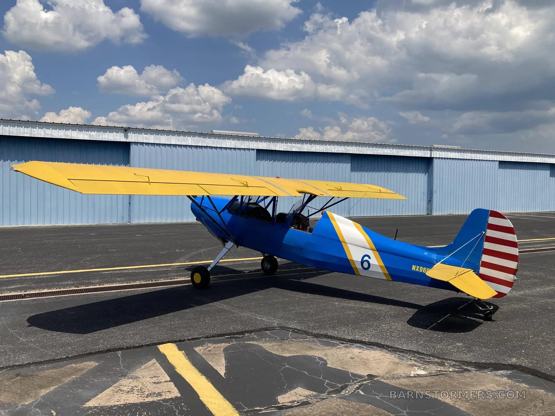 A blue and yellow biplane with '6' on the side, parked on an asphalt runway with hangars in the background.