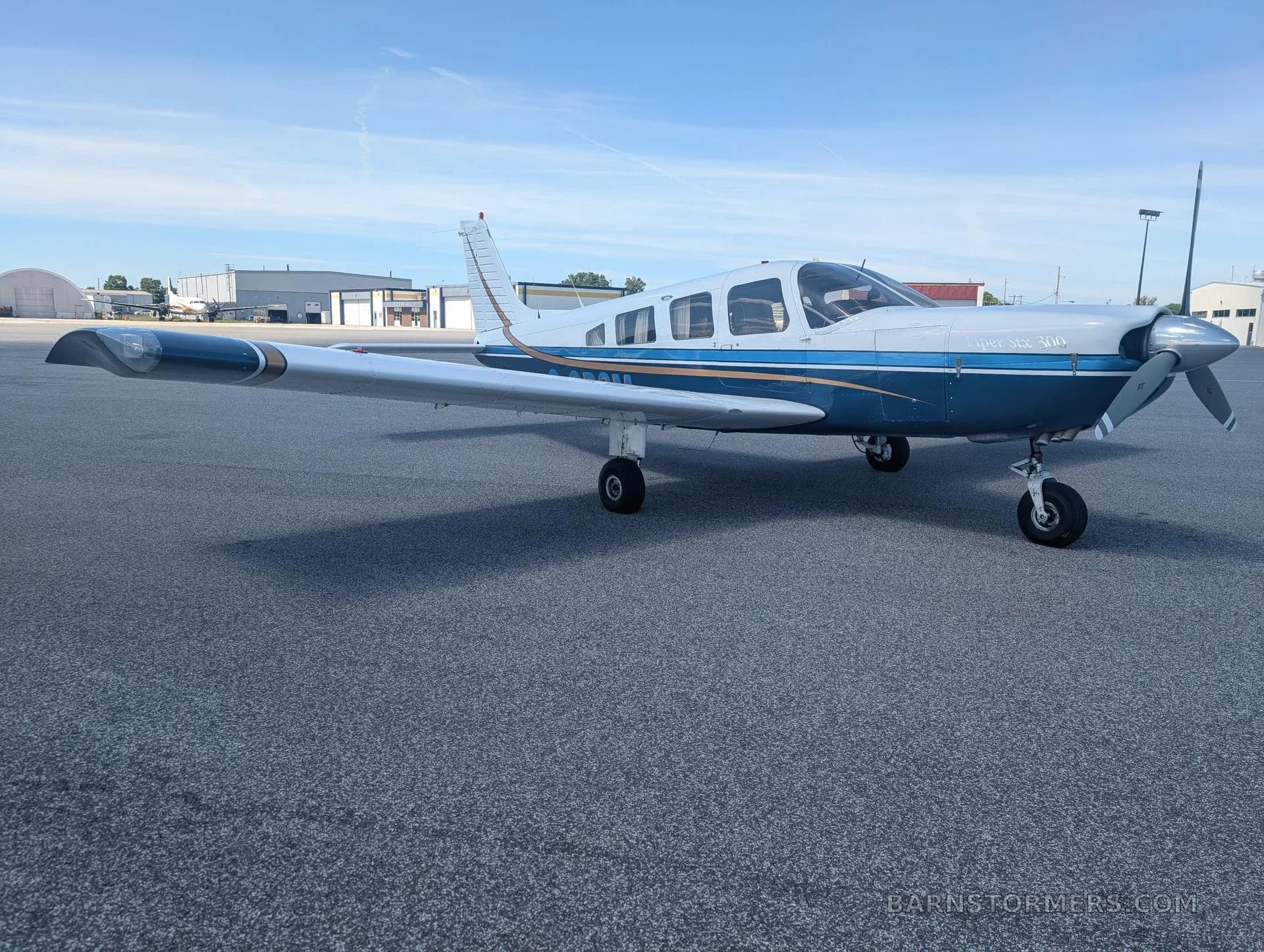 A white and blue Piper Archer 200 aircraft with gold stripes is parked on an asphalt tarmac under a clear blue sky.