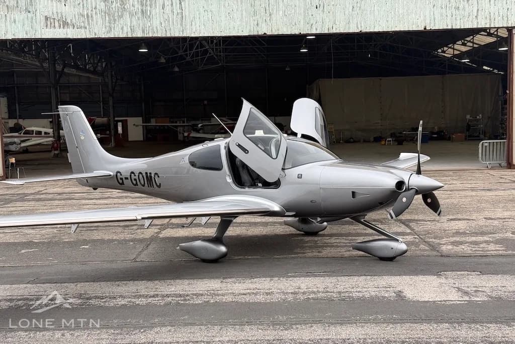 A silver Cirrus SR22 aircraft with its doors open, parked on an airfield in front of a hangar.