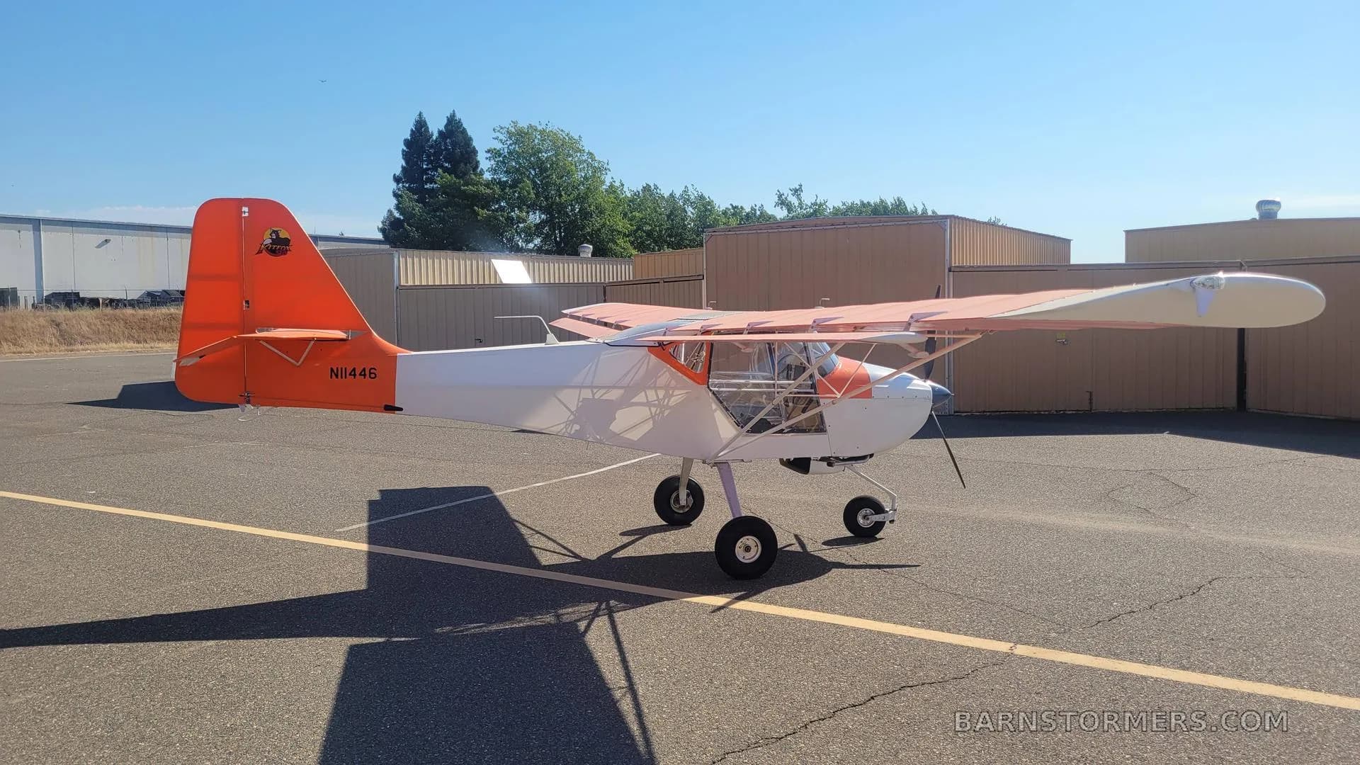 A white and orange high-wing aircraft with tail number N11446, parked on an asphalt apron with hangars and trees in the background.