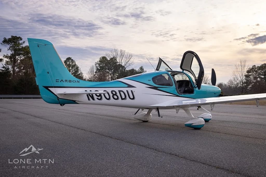 A rear-side view of the teal and white Cirrus SR22 aircraft, N908DU, with its doors open, on an airfield.