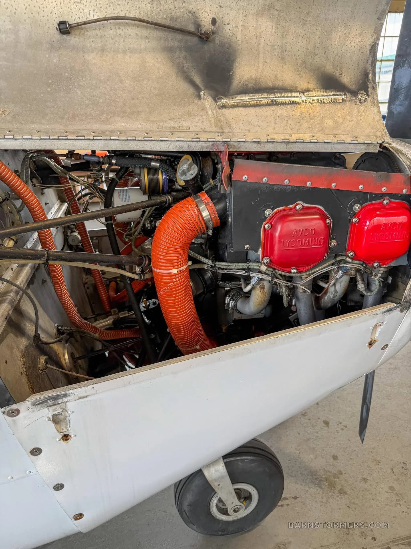 The open engine compartment of a white aircraft, revealing the red AVCO Lycoming engine and orange hoses.