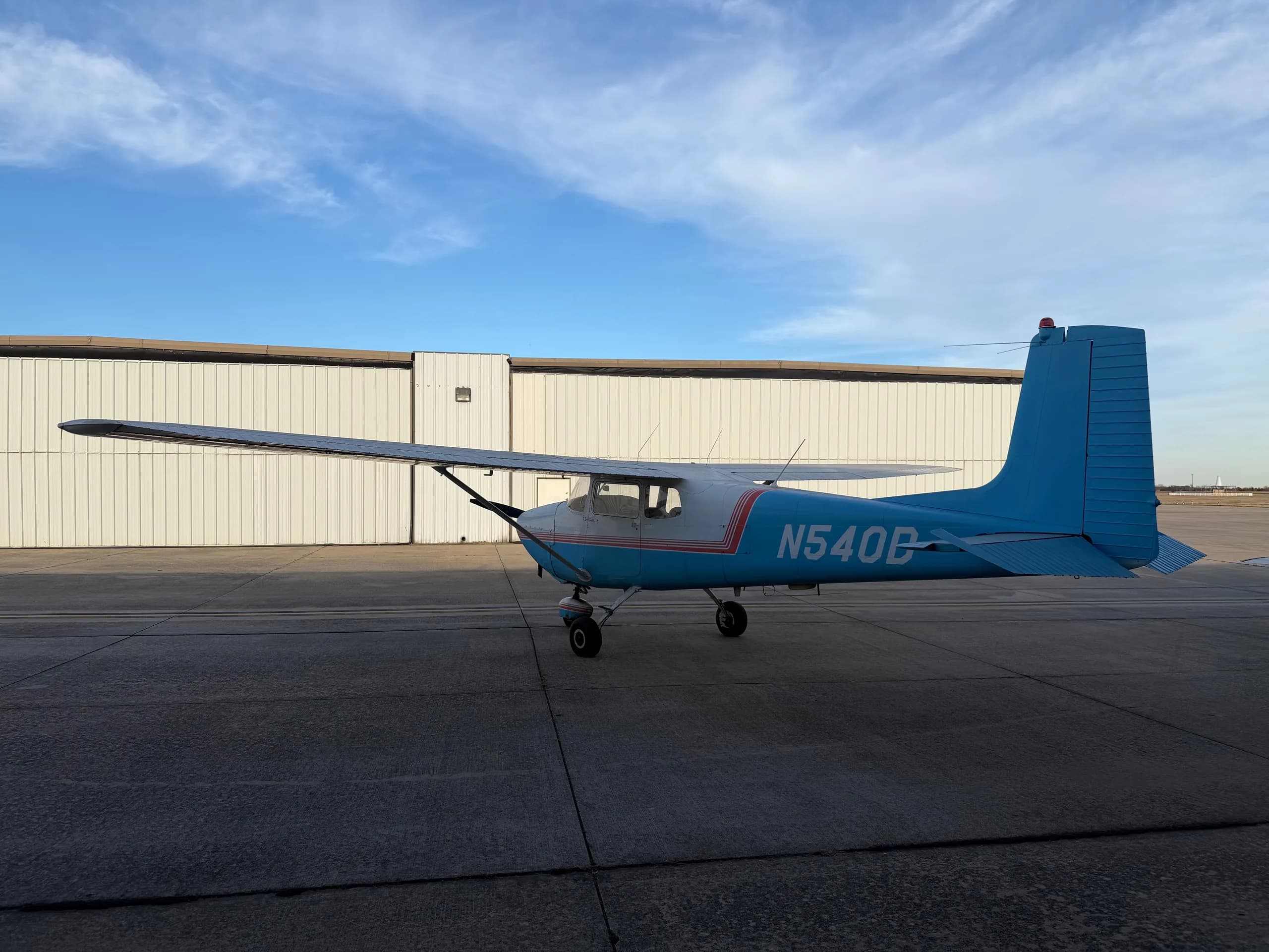 A right-rear view of a white and light blue Cessna 172, registration N540D, parked on an airfield with hangars in the background.