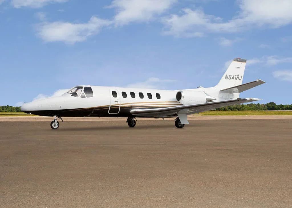 A white and black Cessna Citation business jet N941RJ with gold stripes is parked on a tarmac under a partly cloudy sky, viewed from the front-left.