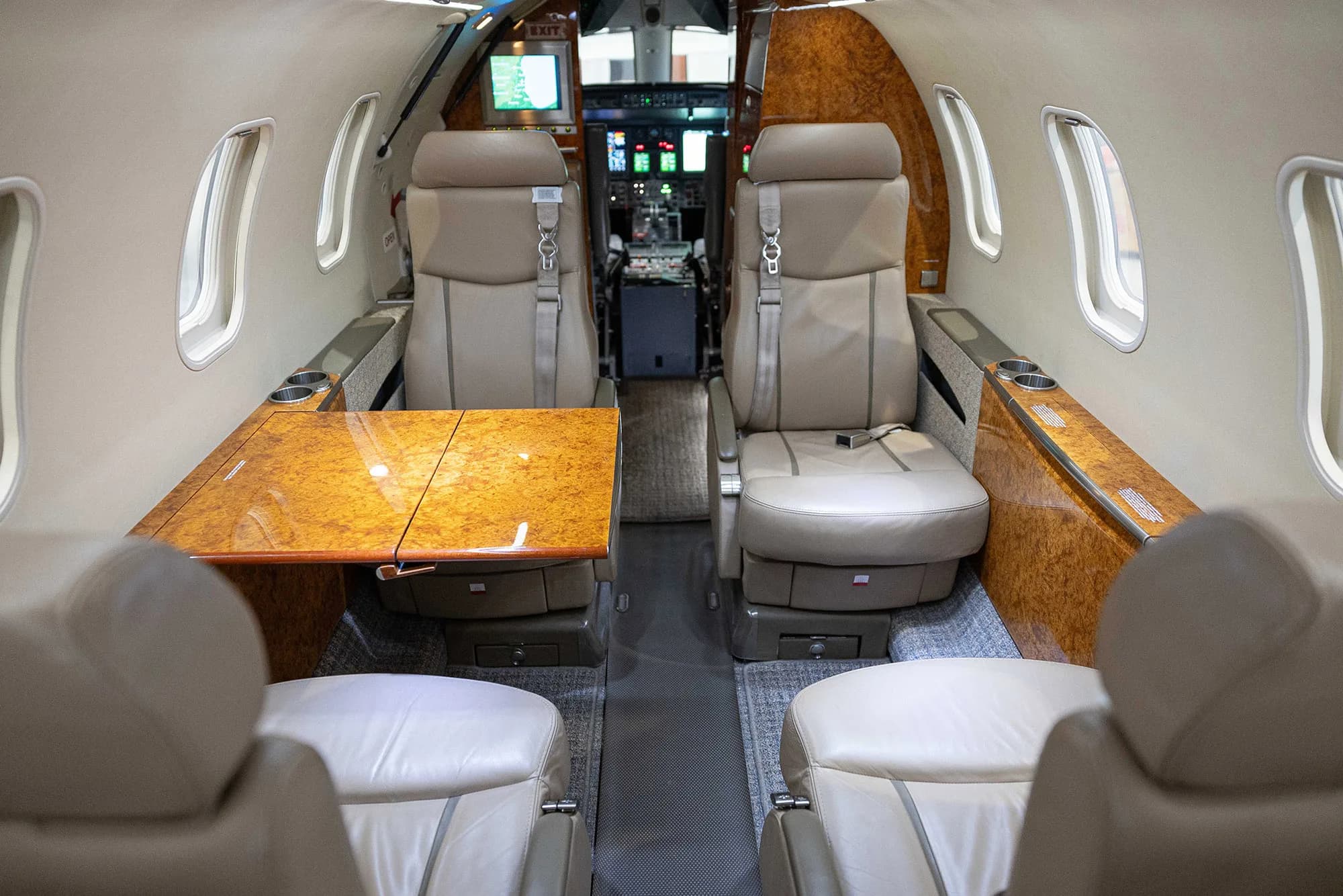 The luxurious interior cabin of a private jet with tan leather seats, a polished wood fold-out table, and the cockpit visible in the distance.