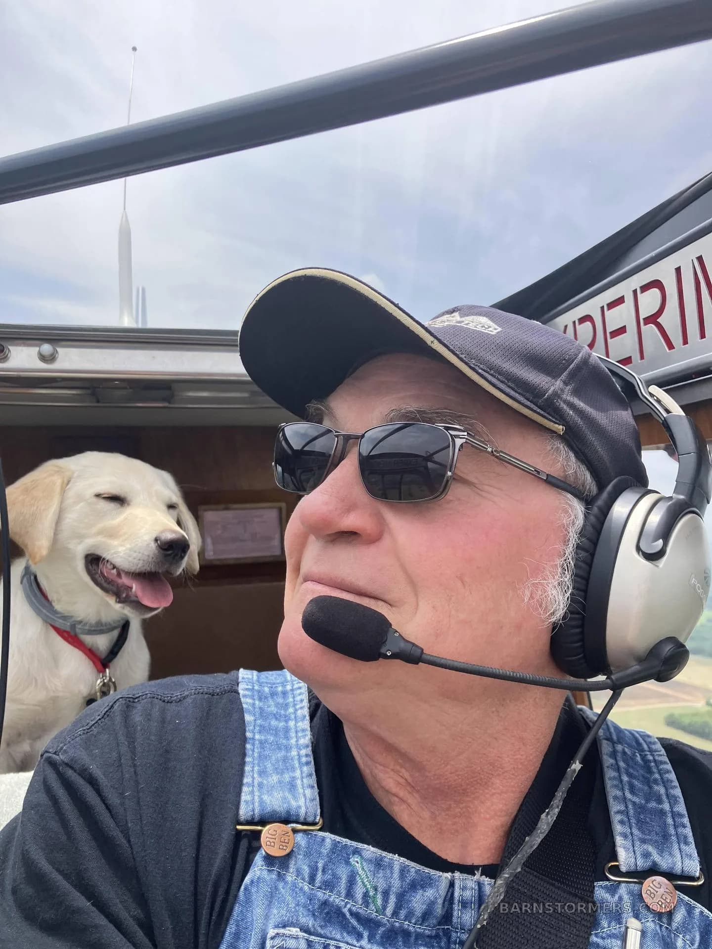 A pilot wearing a headset and sunglasses, with a happy yellow labrador puppy looking out the window from inside an aircraft.
