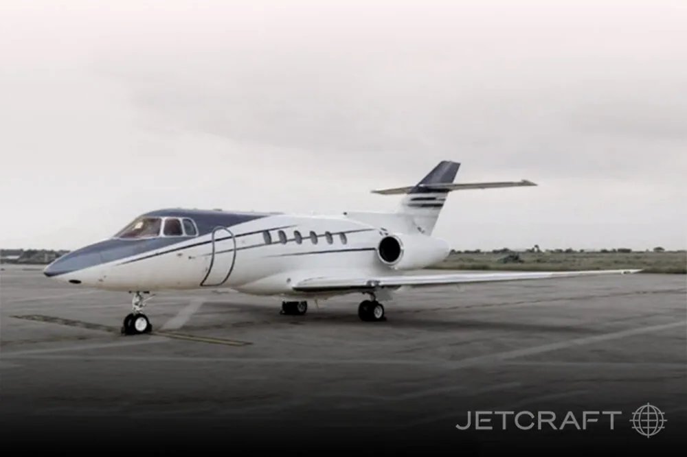 A white and blue business jet, possibly a Hawker, parked on an asphalt tarmac under an overcast sky.