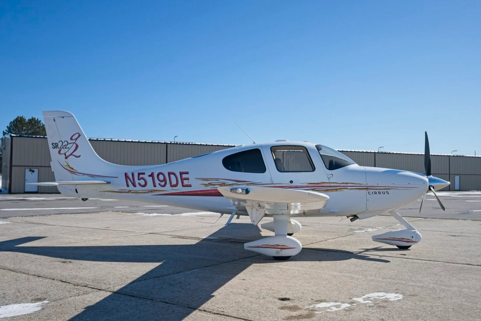 A white Cirrus SR22 aircraft with red and gold decals, registration N519DE, parked on a tarmac with buildings in the background.