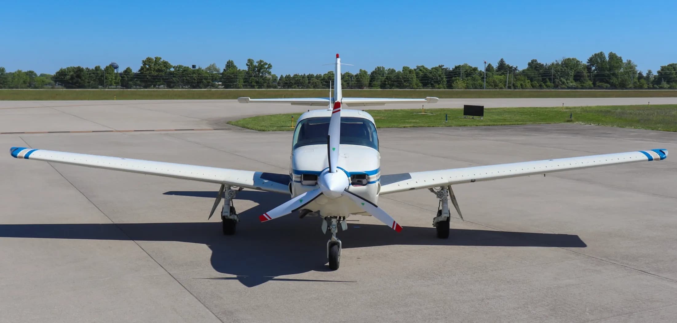 Front view of a white single-engine propeller aircraft with blue and tan stripes, parked on an airfield.