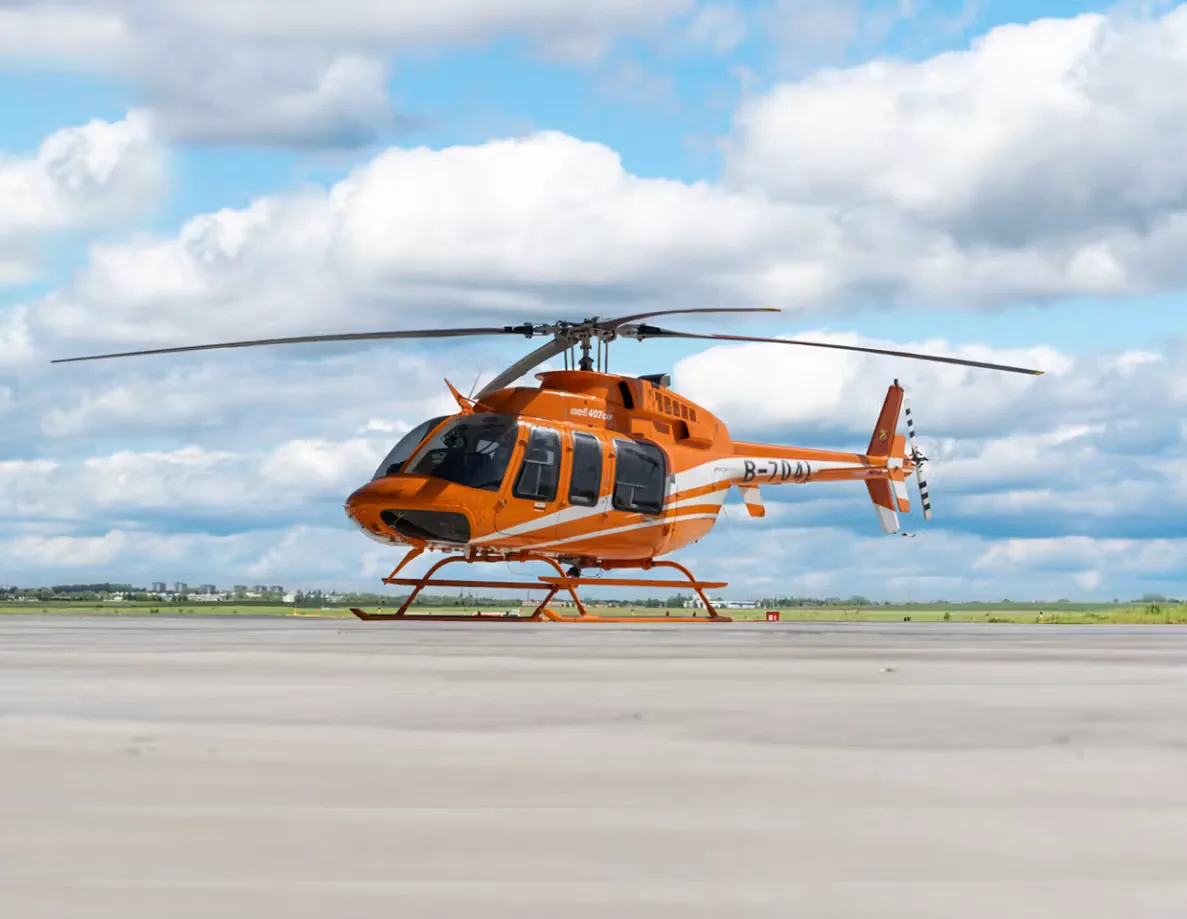 An orange and white Bell 407GXi helicopter is parked on the tarmac under a cloudy sky.