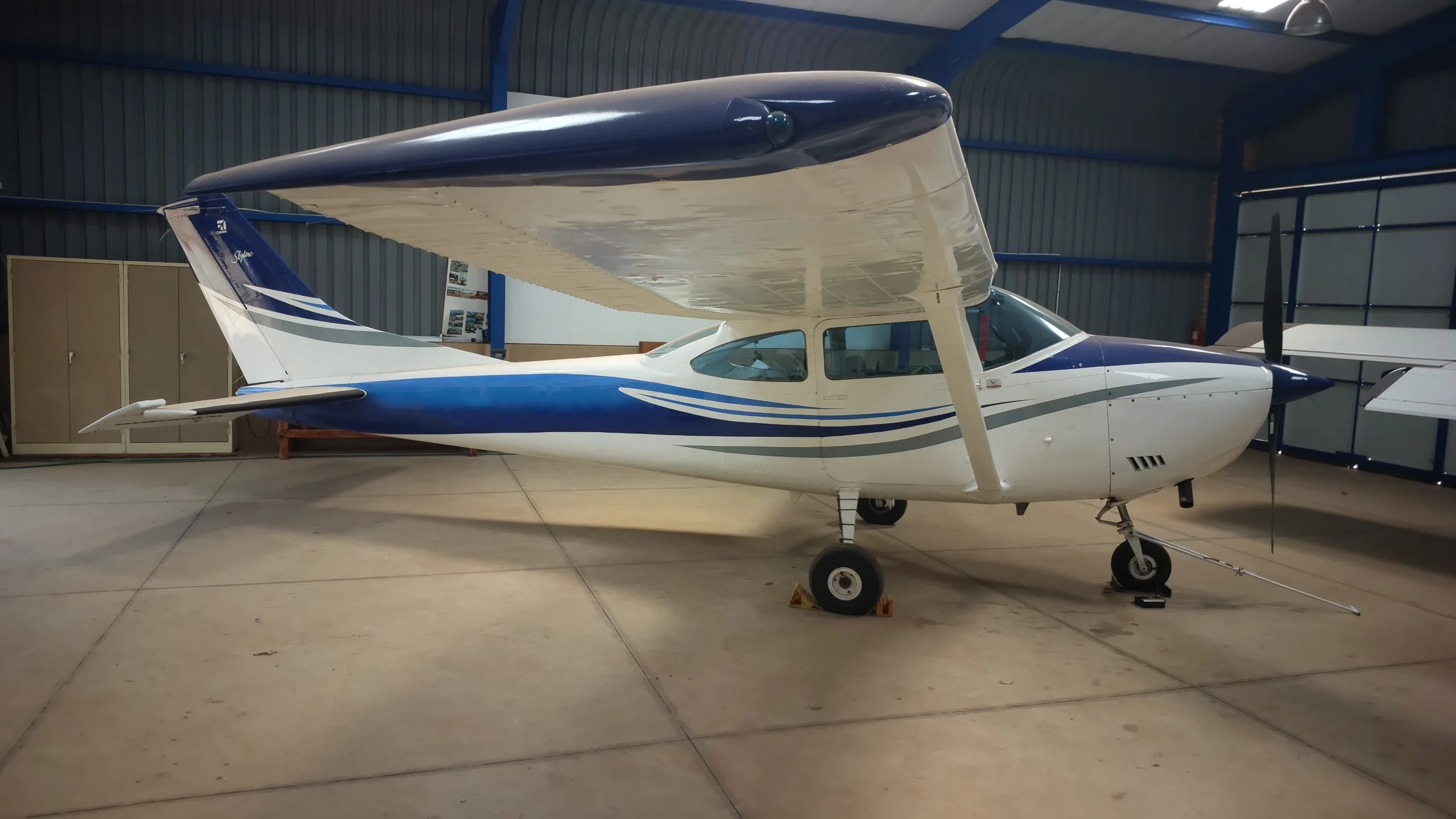 A white and blue Cessna 182 Skylane aircraft is parked inside a hangar.
