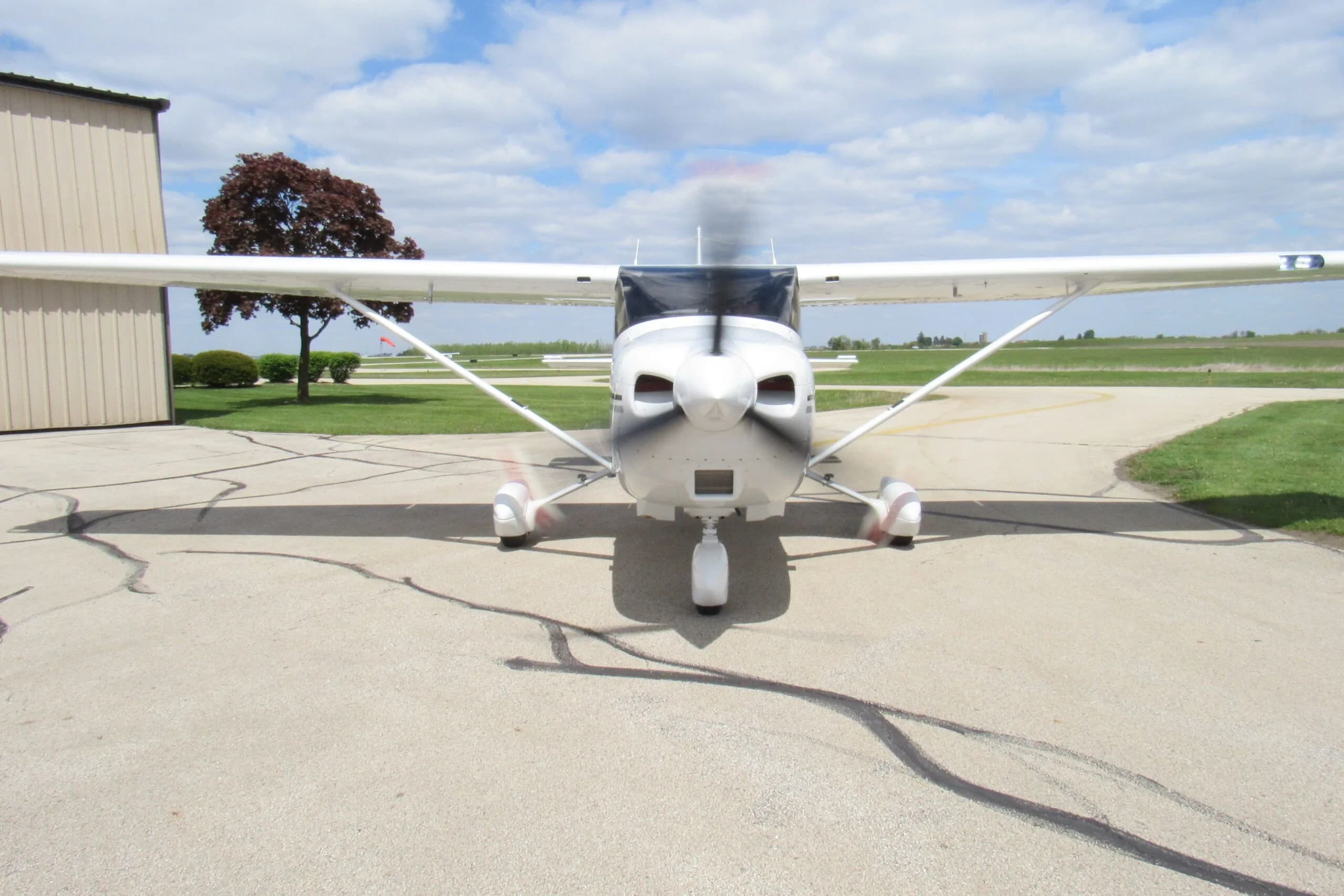 A front view of a white single-engine propeller aircraft with its propeller spinning on an airport tarmac.