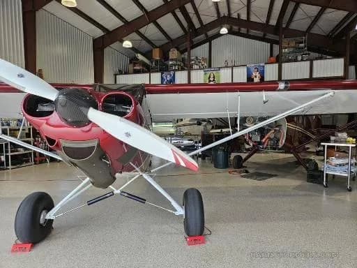 A red and white CubCrafters aircraft with a smiling face painted on the cowling, parked inside a hangar.