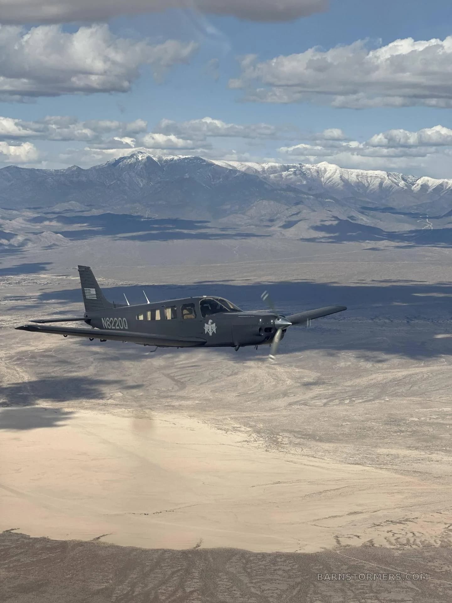 A gray single-engine propeller aircraft, N8220Q, flying over a desert landscape with snow-capped mountains in the background under a partly cloudy sky.