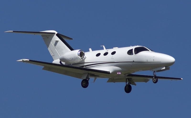 A white jet aircraft with black accents is in flight against a clear blue sky.