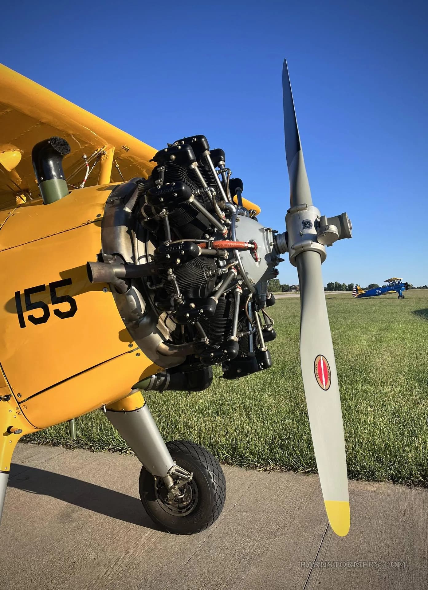 A close-up of the radial engine and propeller of a yellow biplane, with another aircraft visible in the grassy background under a clear blue sky.
