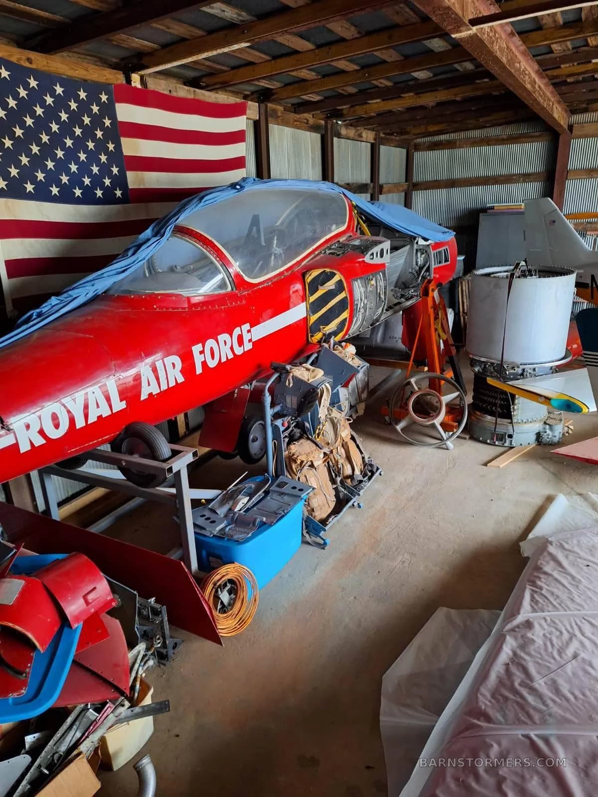 A red jet aircraft, partially covered and disassembled, sits in a hangar with an American flag hanging in the background.