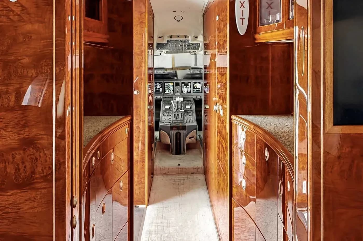 A narrow hallway with highly polished wood cabinetry leads towards the cockpit of a business jet, with the instrument panel visible in the distance.