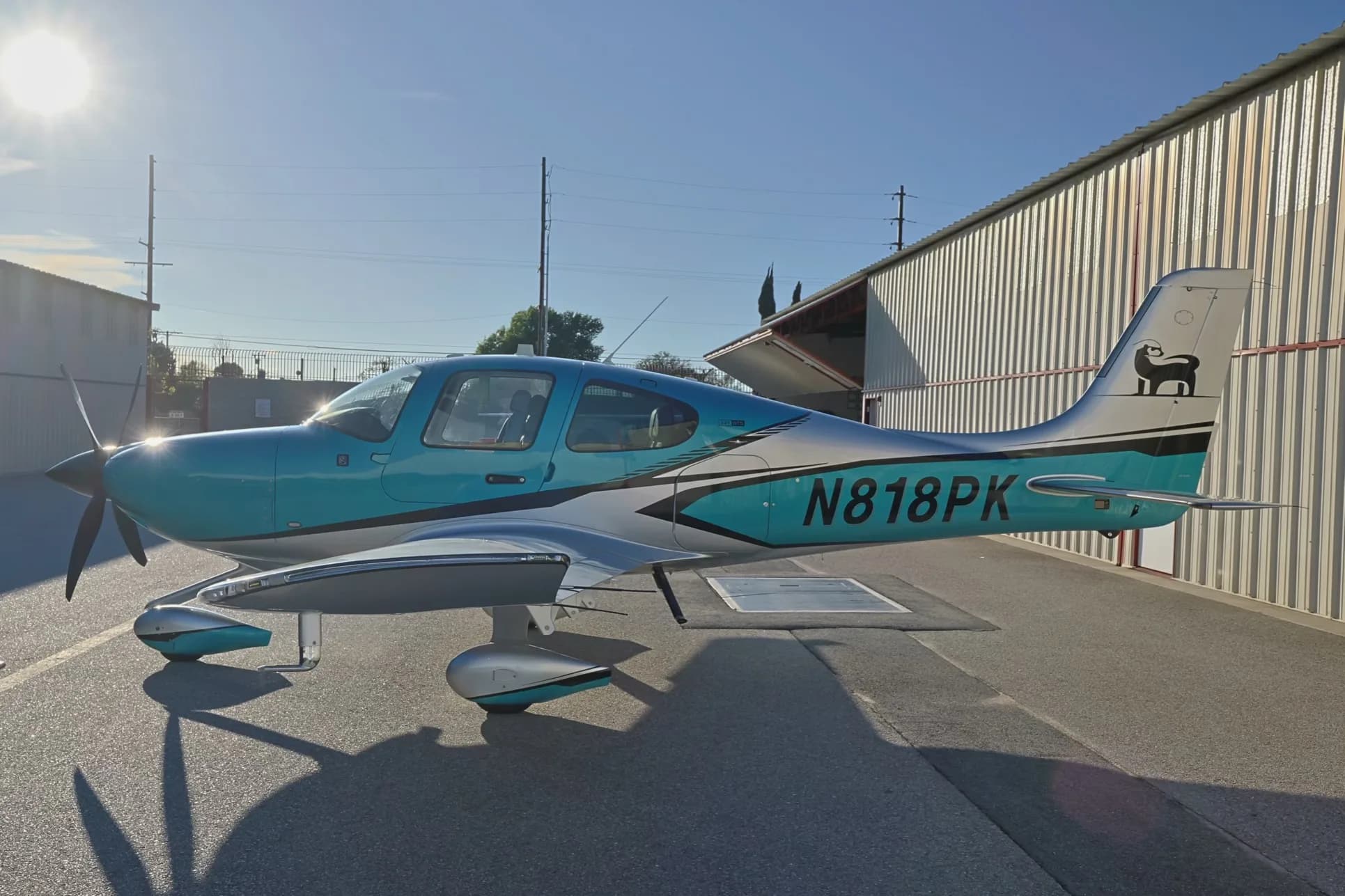 A side view of a teal and silver Cirrus SR22T GTS aircraft (N818PK) parked on an airfield with sun glare.