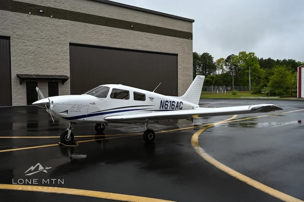 A white single-engine aircraft, N616AC, with blue stripes is parked on a wet tarmac with trees in the background.