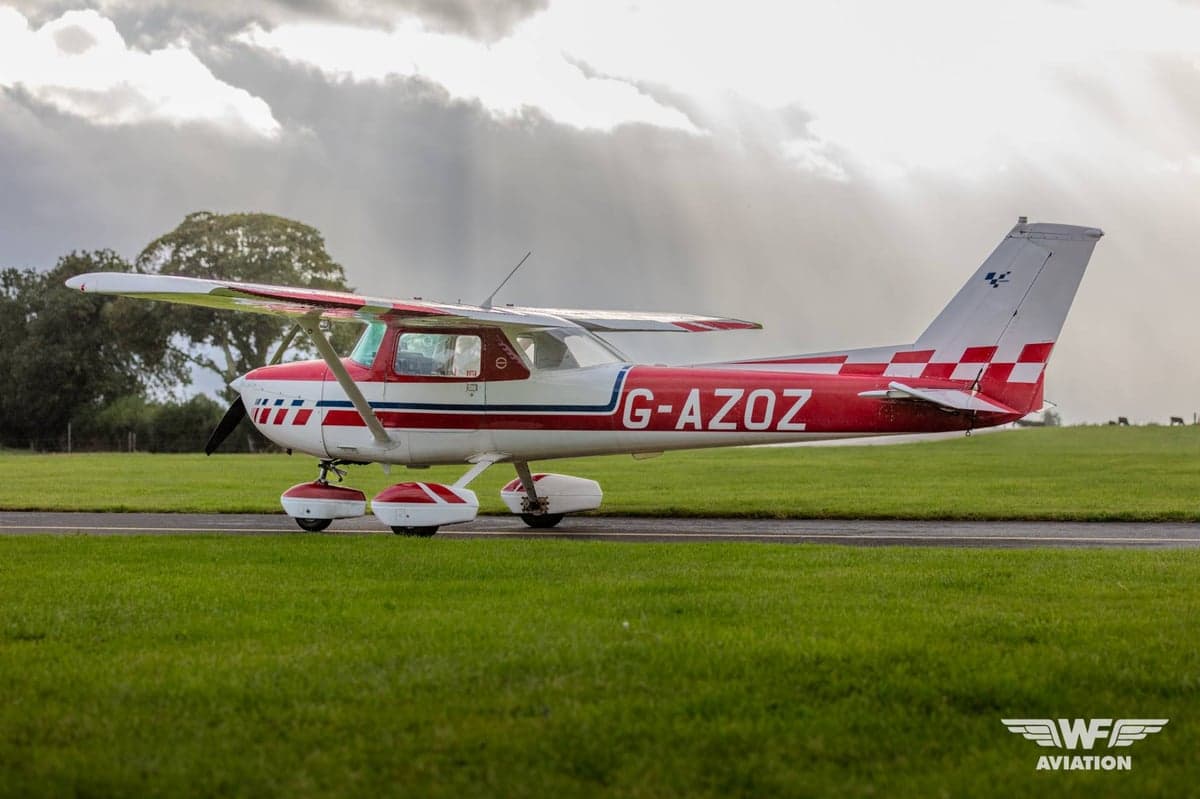 A red and white Cessna 150 Aerobat (G-AZOZ) with a checkered tail, parked on a tarmac with sun rays breaking through clouds.