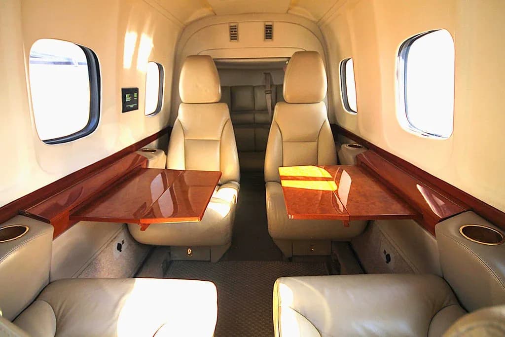 The aircraft cabin interior features two rows of beige leather seats with fold-out wooden tables.
