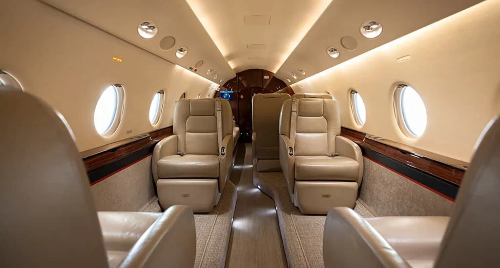 The luxurious interior cabin of a business jet, featuring tan leather seats arranged along a central aisle.