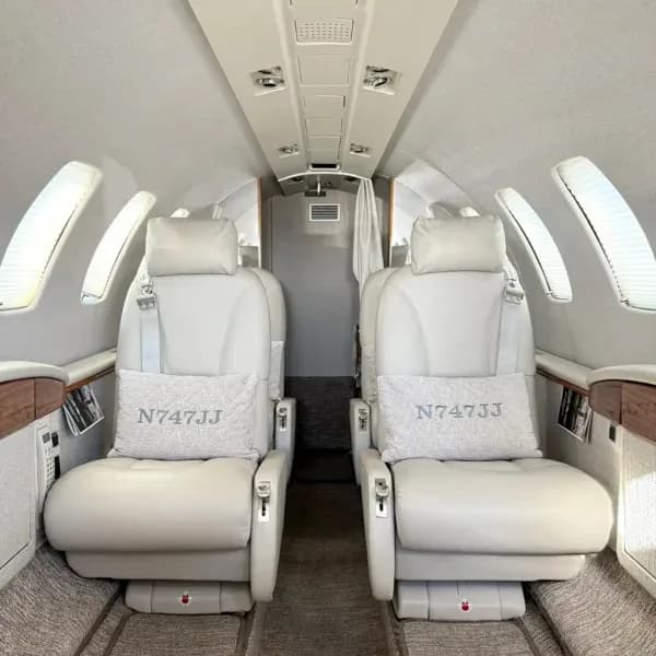 Interior view of a private jet cabin with light-colored leather seats and pillows embroidered with 'N747JJ'.