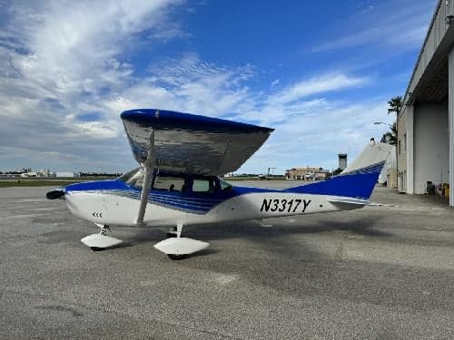 A Cessna 182 Skylane aircraft with a blue and white paint scheme is parked on the tarmac.