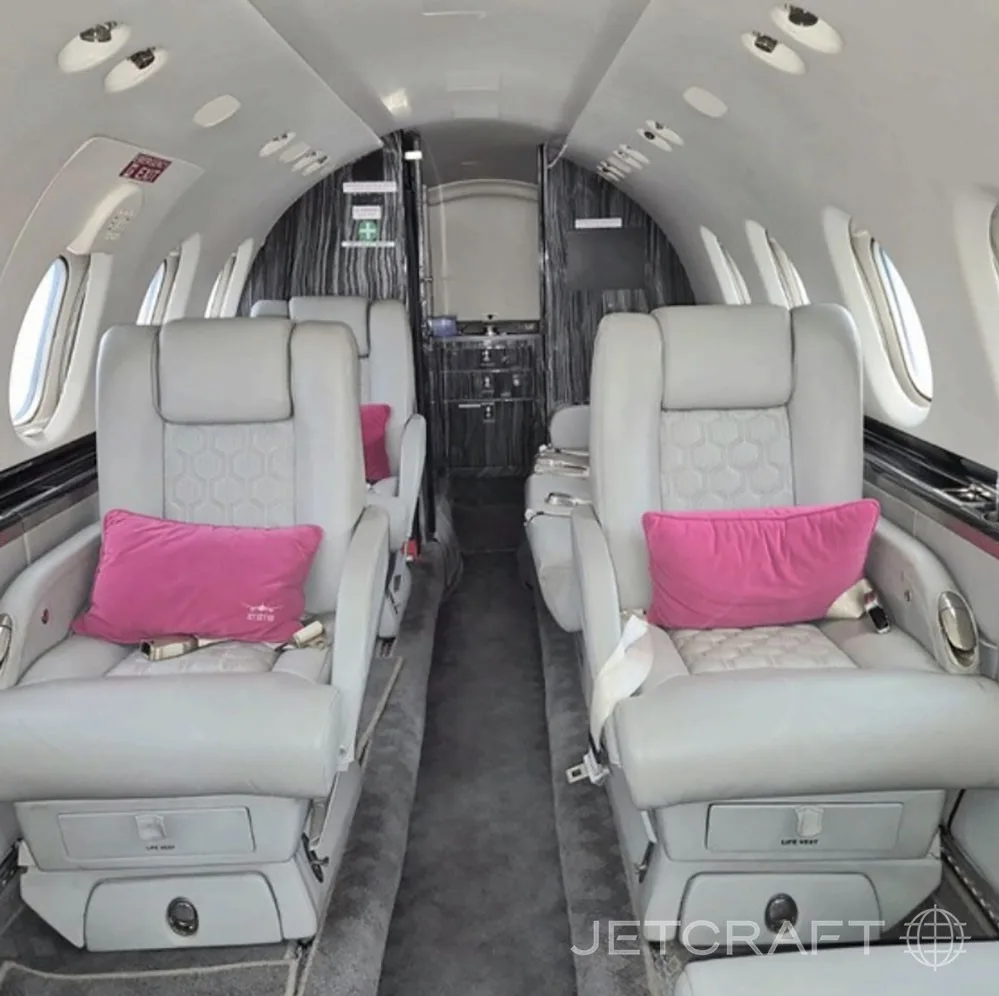 The interior cabin of a business jet with light grey leather seats, pink pillows, and a dark grey aisle carpet.