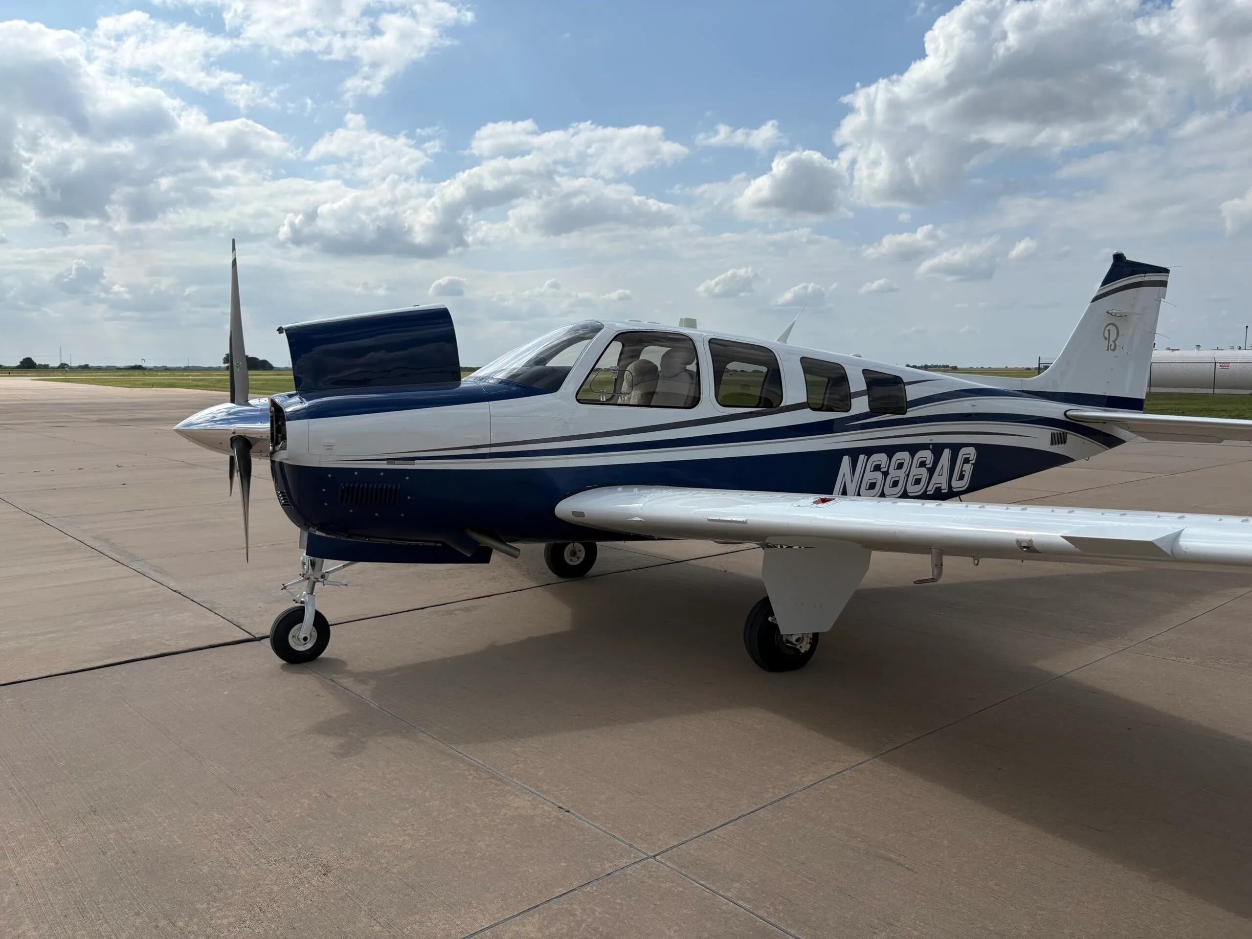 A side view of the white and blue Beechcraft Bonanza N686AG with its engine cowling open, parked on an airfield.
