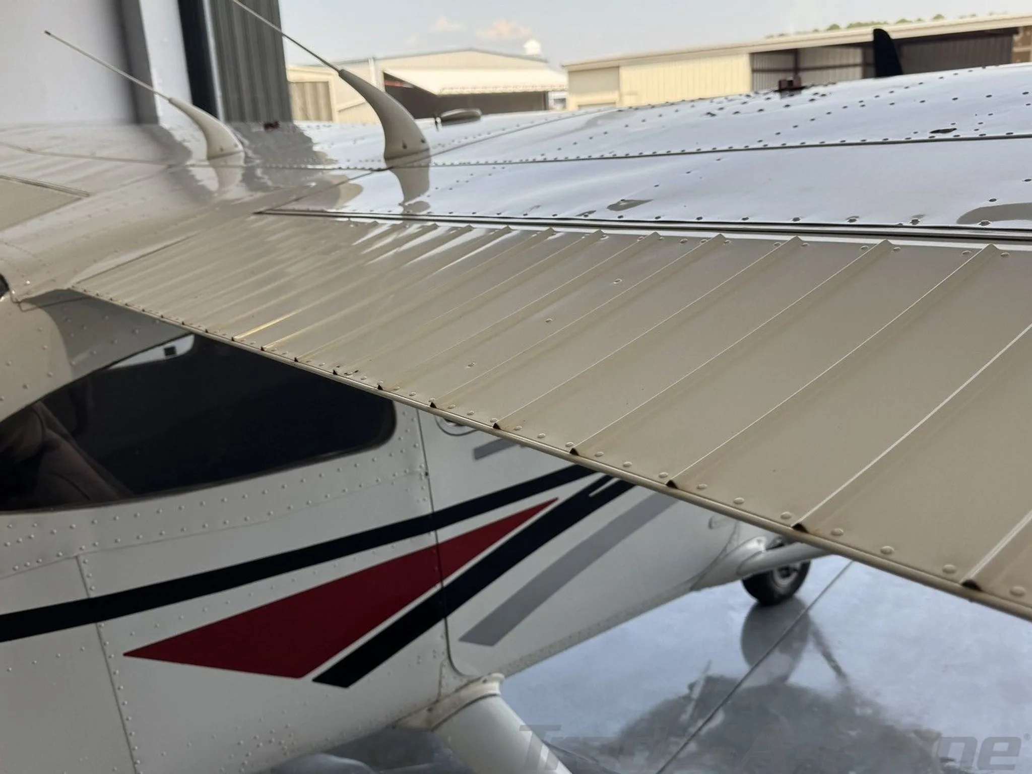 A close-up view of the wing and upper fuselage of the white Cessna 182 Skylane, showing rivets and antennas, inside a hangar.