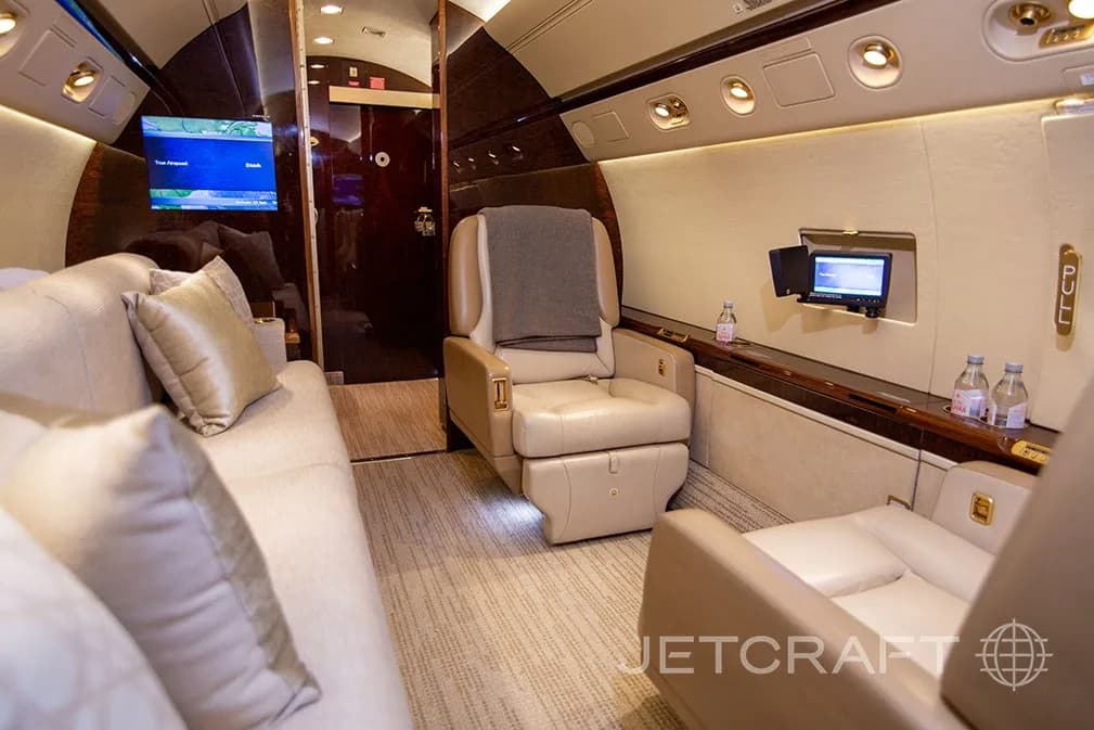 A private jet cabin showing a beige sofa, a reclining seat with a blanket, and a small entertainment screen.