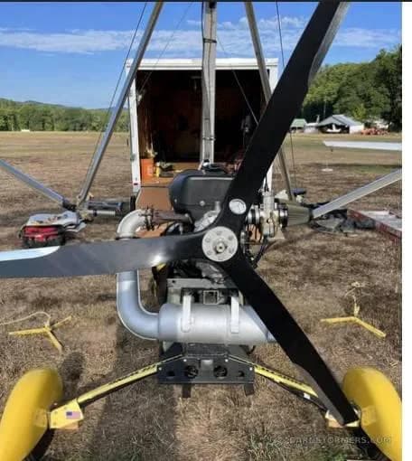 A front view of a yellow ultralight aircraft with a large black propeller, parked on a grassy field with a white trailer in the background.