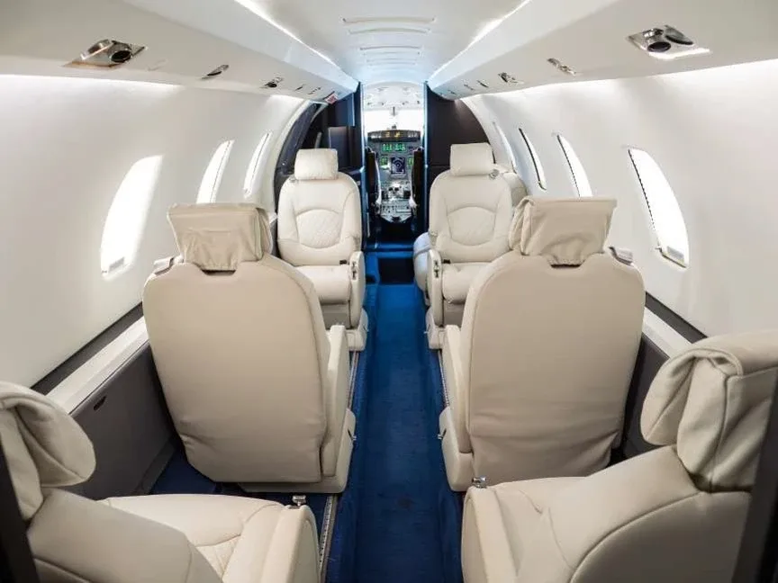 The interior cabin of a business jet, featuring light beige leather seats and a blue carpeted aisle, looking towards the cockpit.