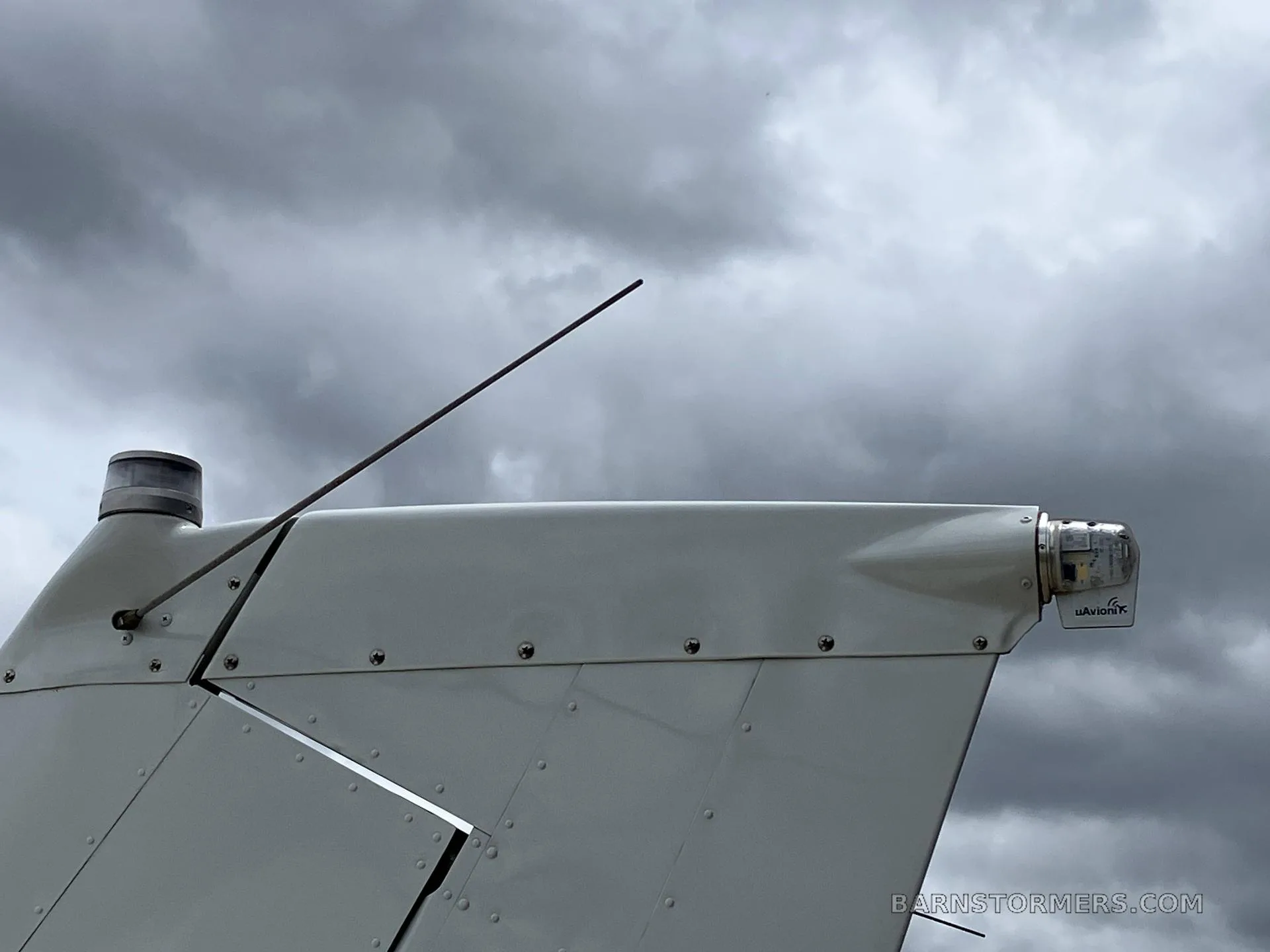 A close-up view of the tail section of a white aircraft, showing an antenna and a navigation light against a cloudy sky.