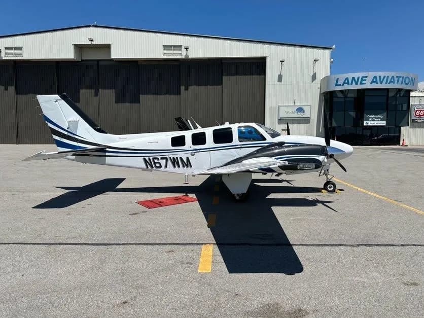 A white and blue twin-engine propeller aircraft, tail number N67WM, is parked on an asphalt ramp in front of a hangar.