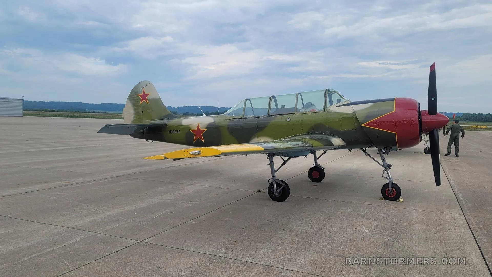 A Yakovlev Yak-52 aircraft in green camouflage with red stars and a red nose, parked on an airfield with a person in the background.