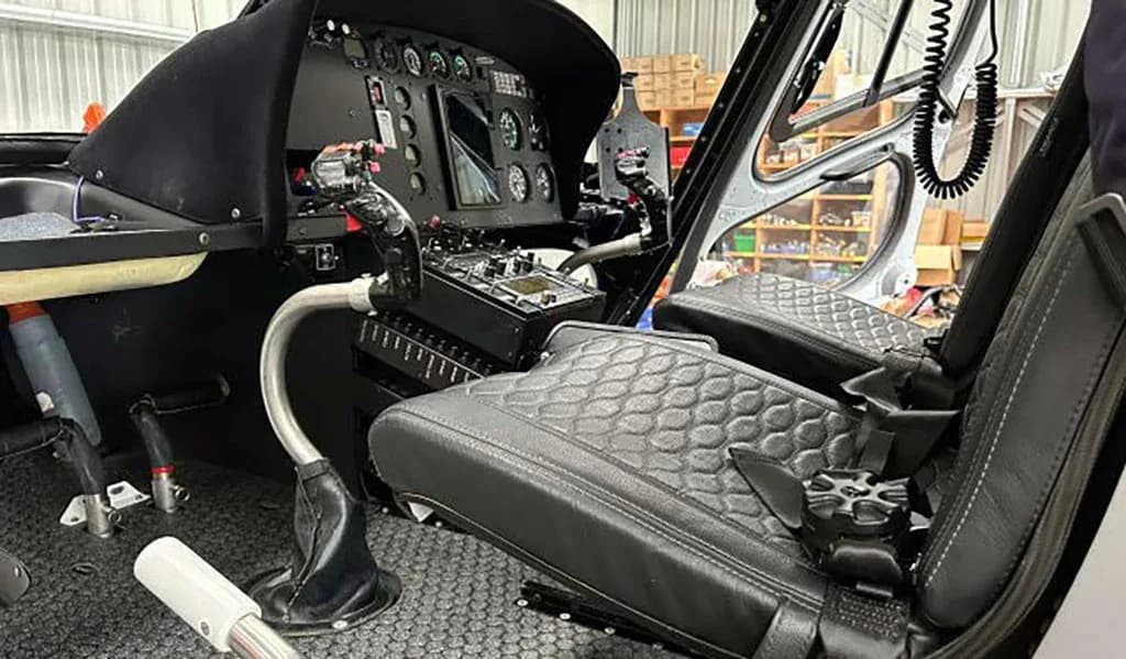 A view of the helicopter cockpit showing the pilot's quilted black seat, flight controls, and instrument panel.