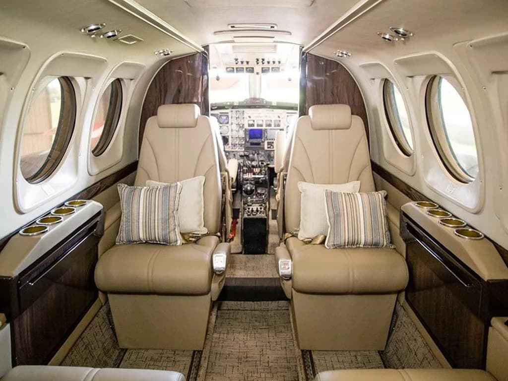 The aircraft cabin interior with two rows of beige leather seats, striped pillows, and a view into the cockpit.